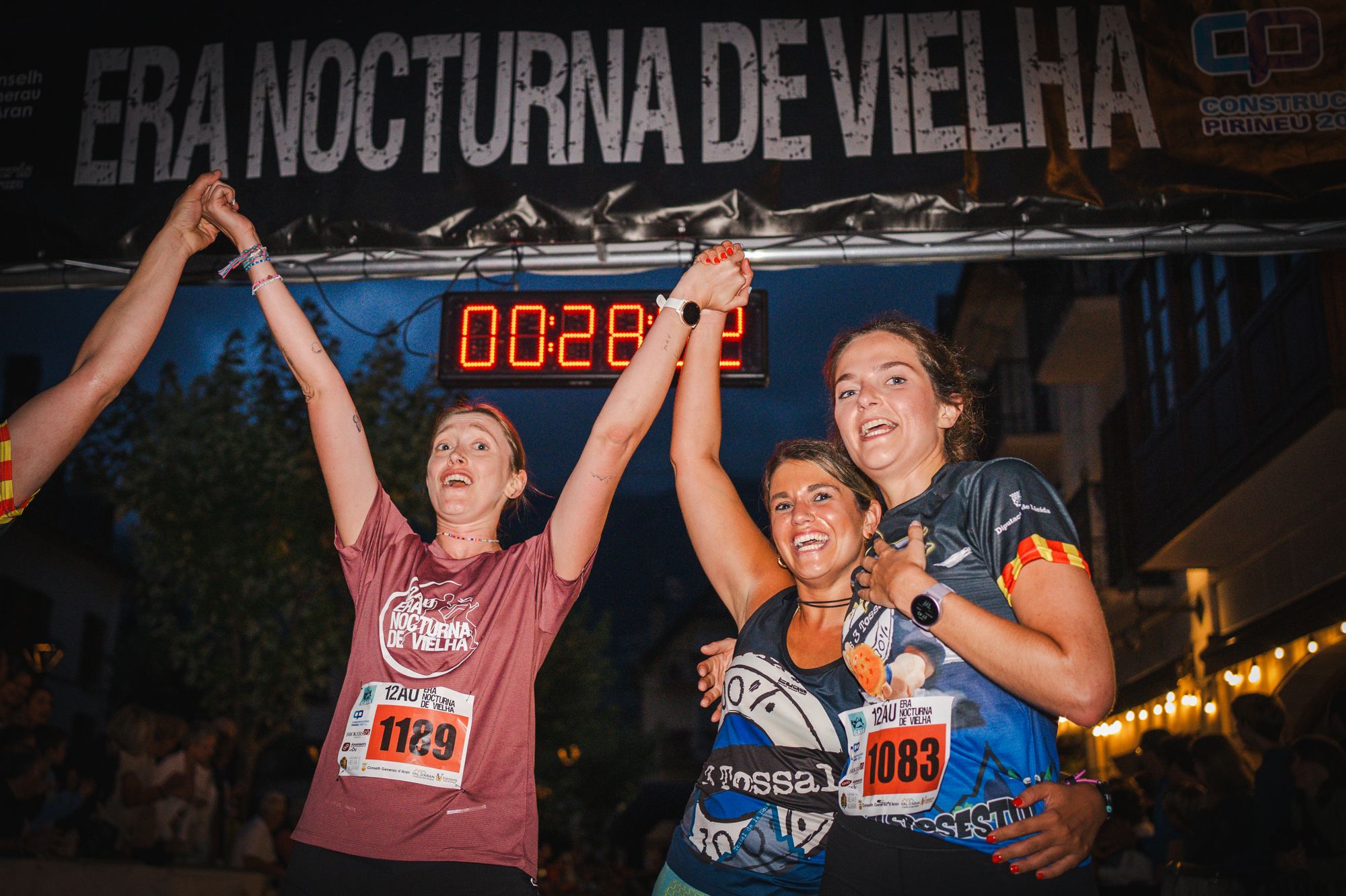 Tres corredoras celebran bajo el reloj de meta, de noche